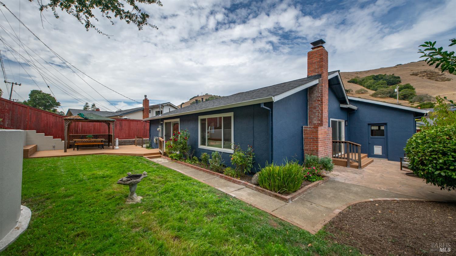 Blue home with brick chimney and landscaped backyard, lawn, patio, and covered gazebo.