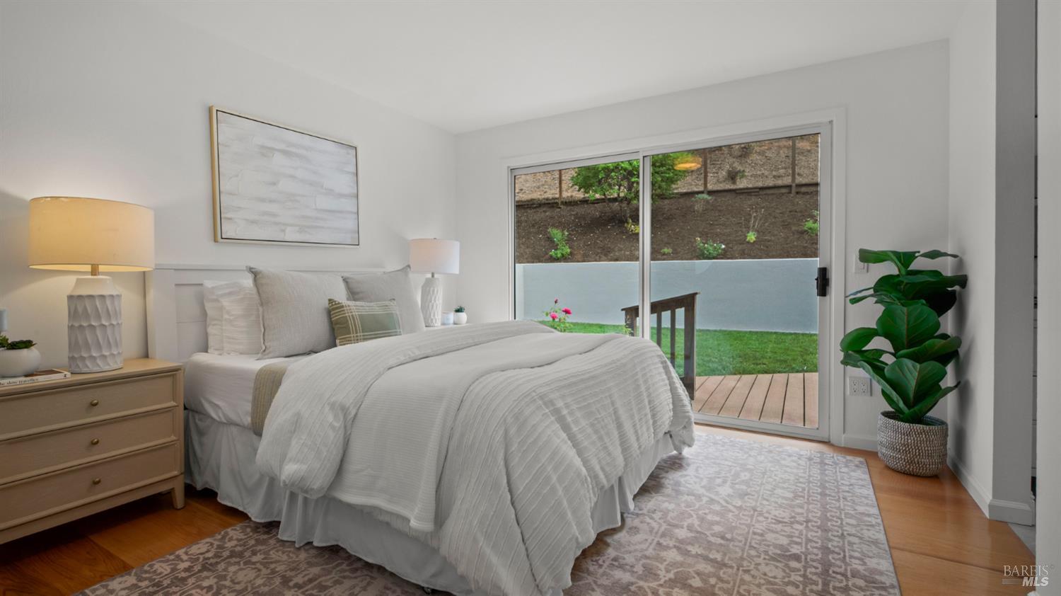 Staged bedroom with a white bed beside sliding glass doors opening onto a deck and garden.