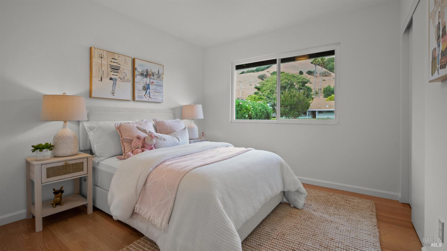 Staged bedroom with a white bed, blush-pink bedding, framed artwork, and a window overlooking a hillside.