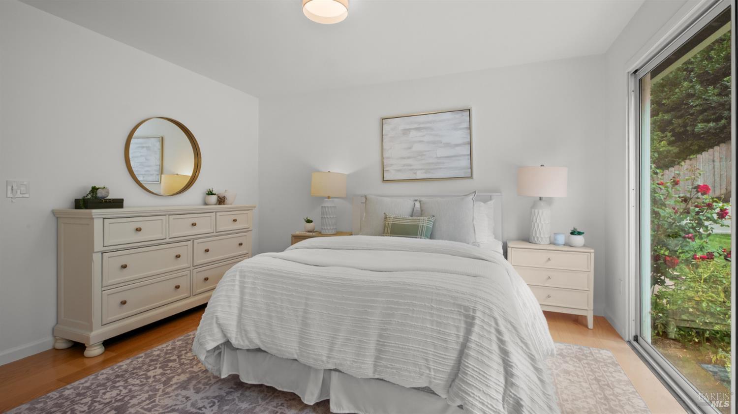 Staged bedroom with a white bed, cream dresser, and sliding glass door overlooking a garden.