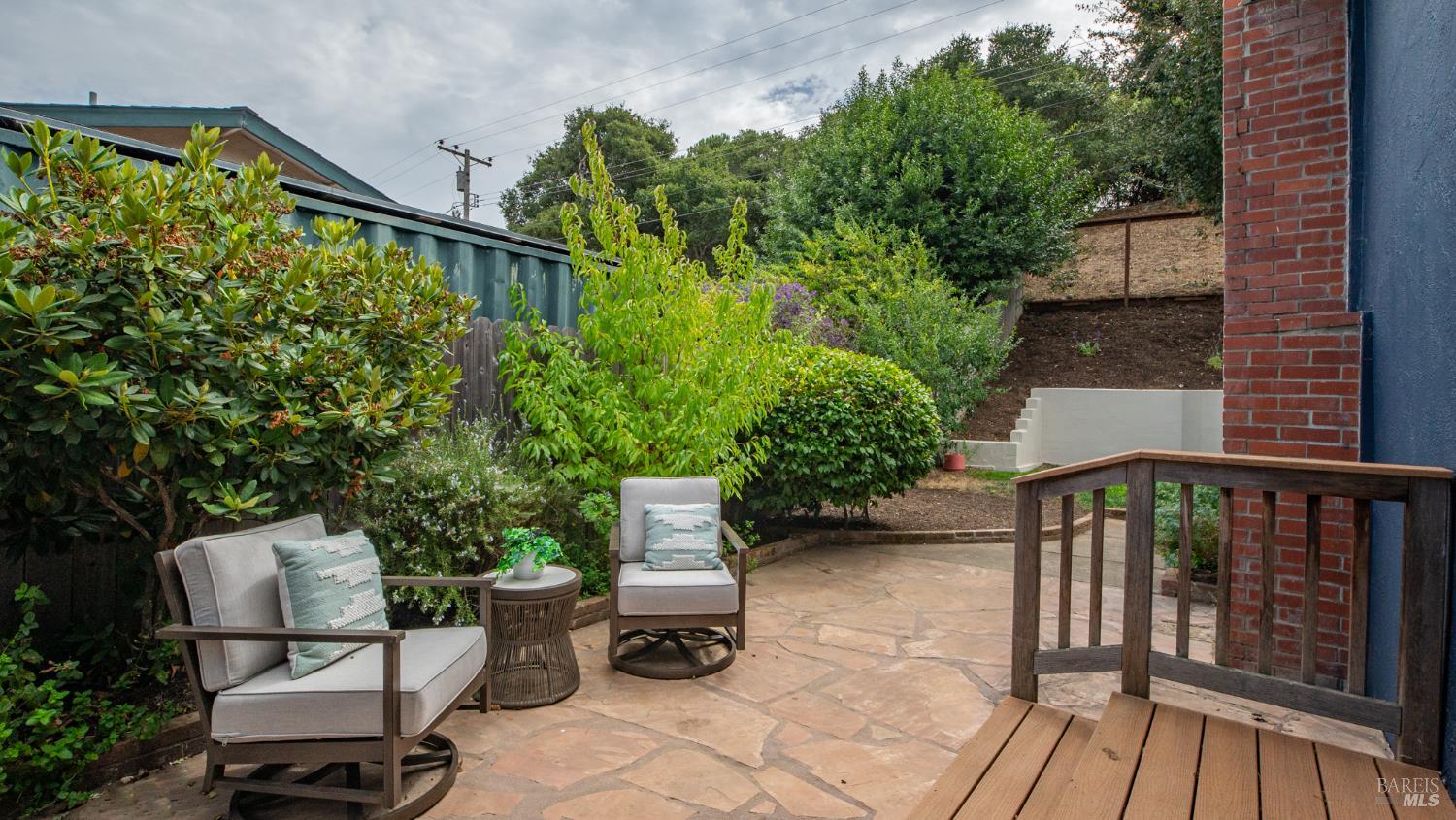 Stone patio with cushioned seating, dense garden shrubs, and a wooden deck railing.