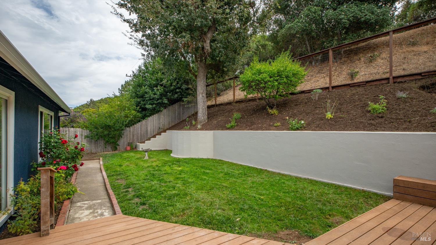 Backyard with a wooden deck, lawn, flower beds, and a curved retaining wall.