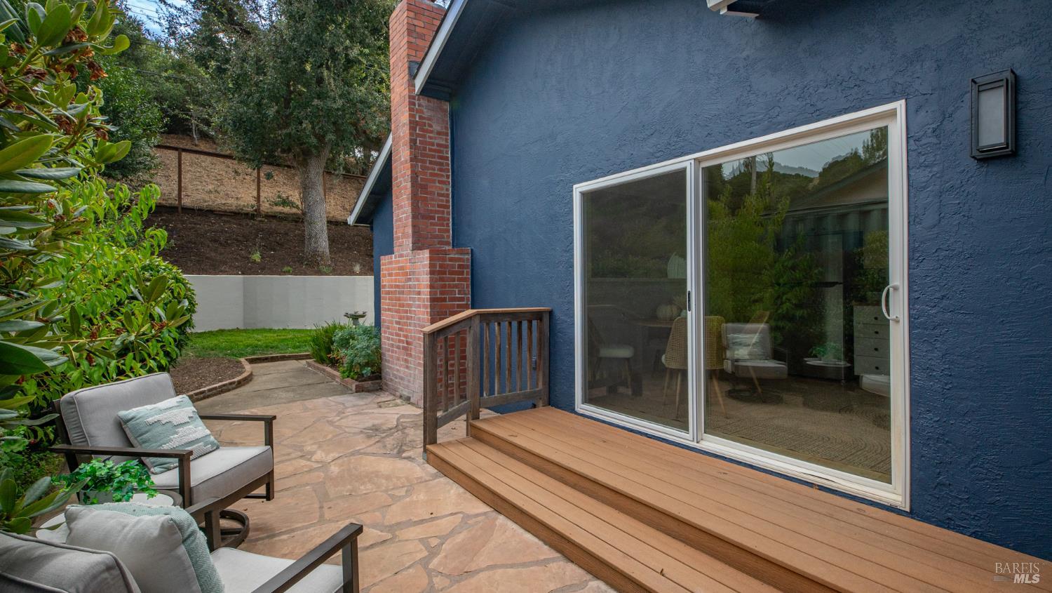 Rear patio with a raised wooden deck, blue exterior wall, and cushioned chairs beside a leafy garden.
