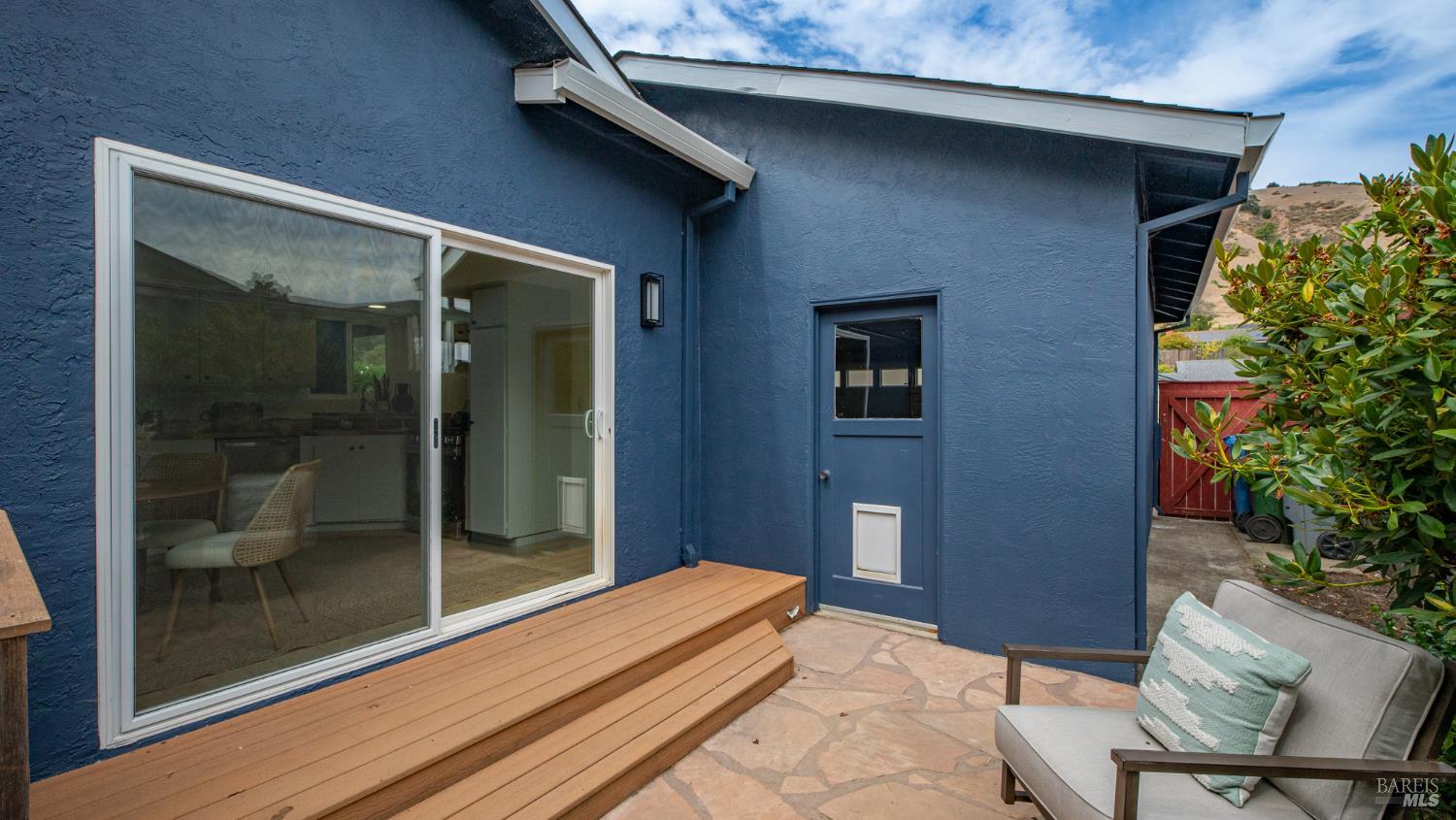 Rear patio with blue stucco siding, wooden steps, stone paving, and a sliding glass door.