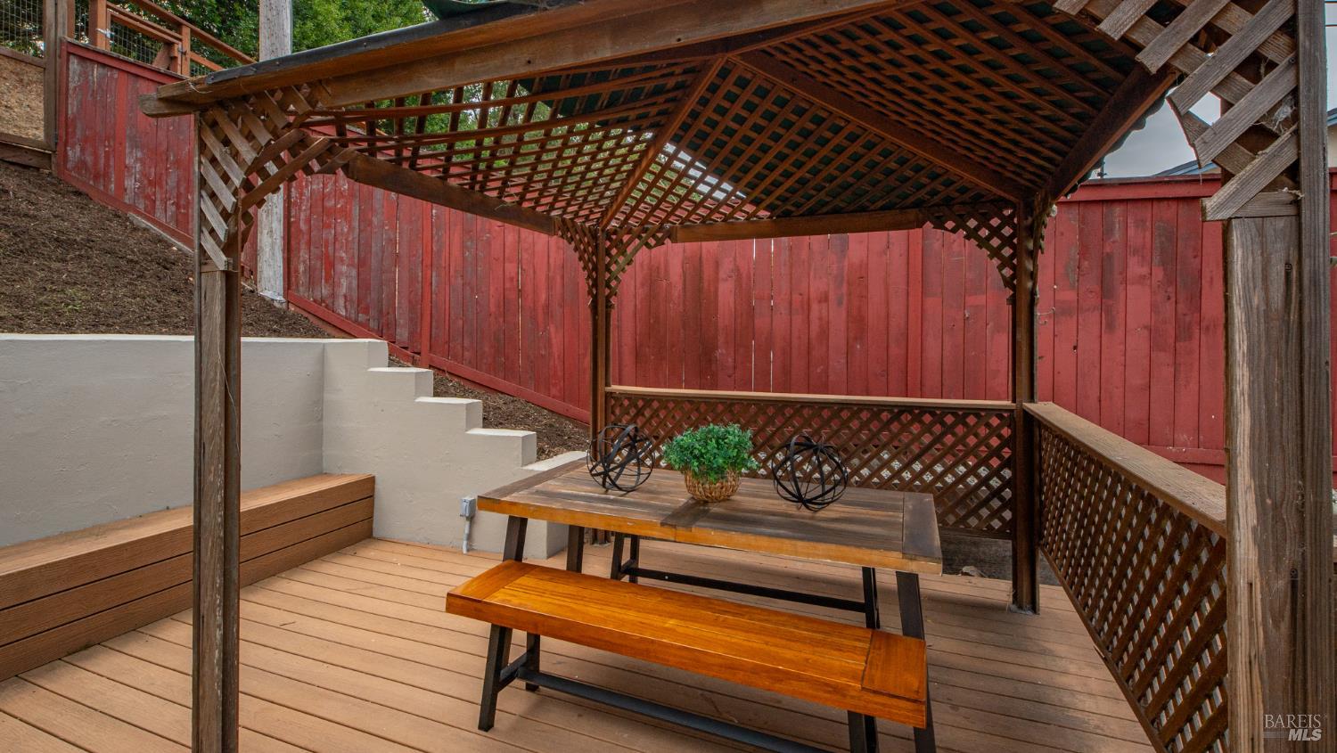 Covered wooden deck with lattice pergola, picnic table, built-in benches, and red fencing.
