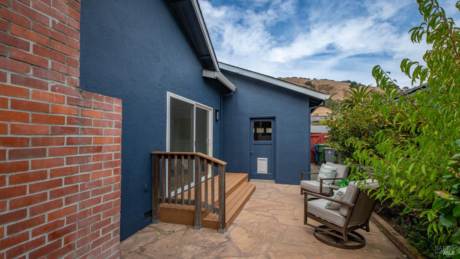 Stone patio beside a blue house, with wooden steps and cushioned seating among shrubs.