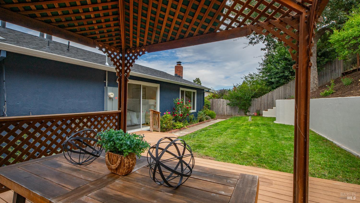 Backyard with a pergola-covered deck, outdoor dining table, green lawn, and flower garden.