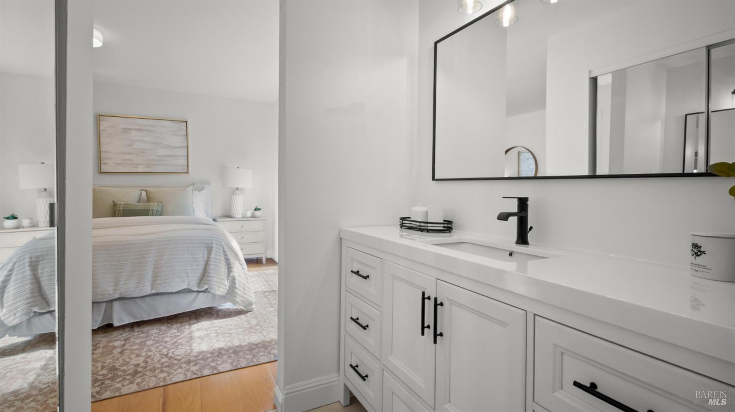 Bathroom vanity with black faucet and large mirror, overlooking an adjoining bedroom.