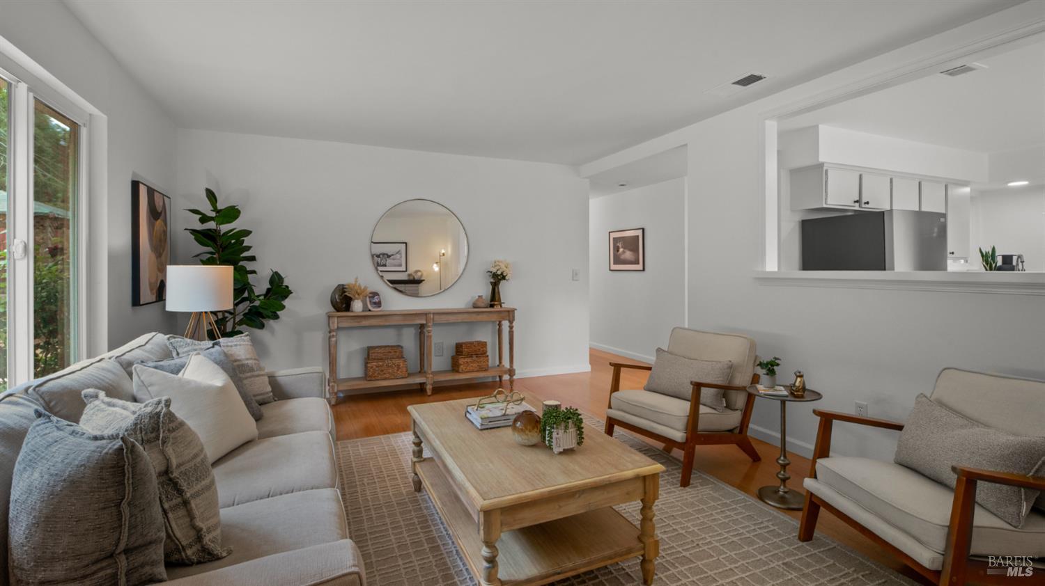Staged living room with a light sofa, armchairs, wooden coffee table, and kitchen opening.