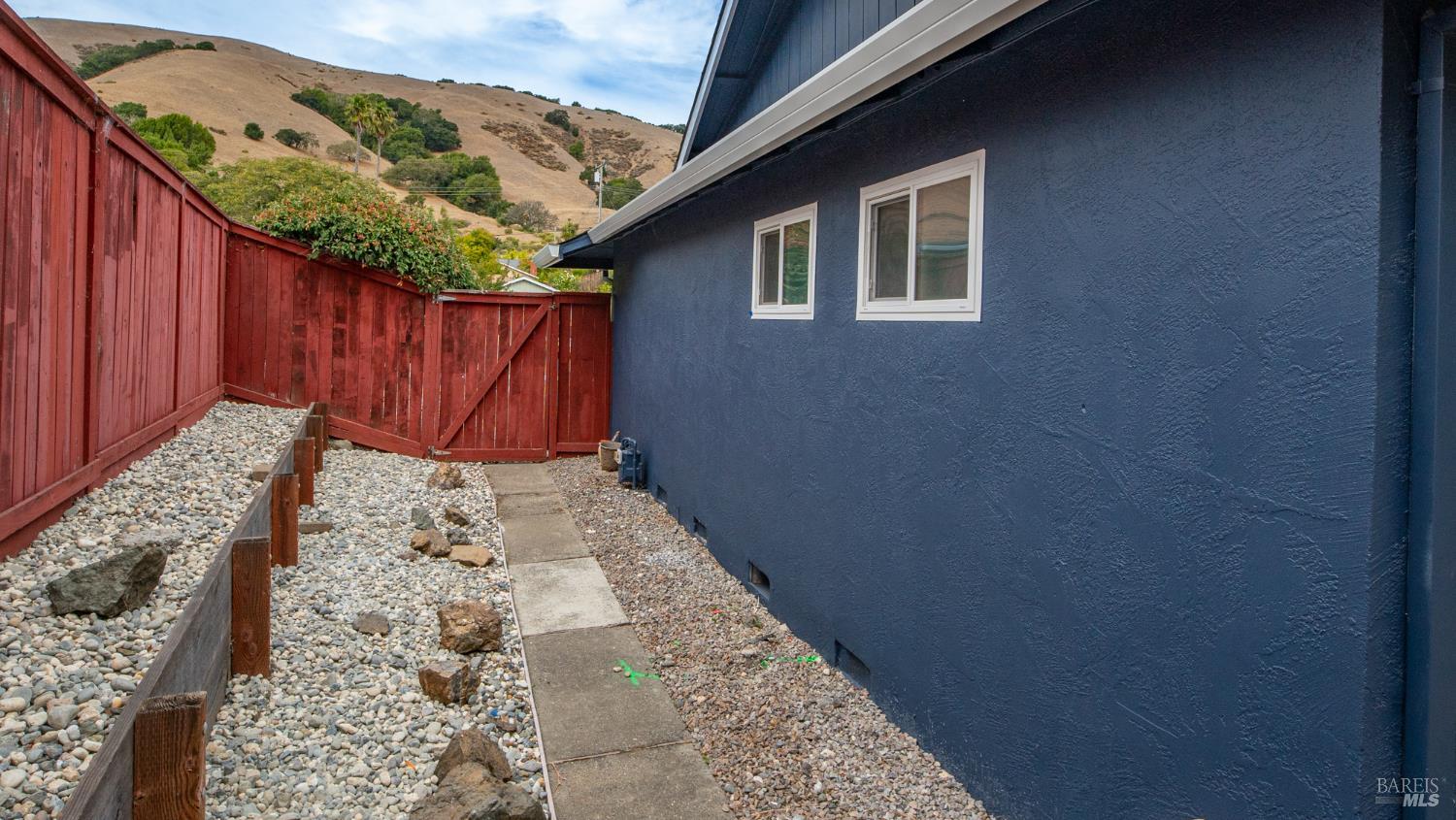 Blue house beside a red fence and rock-covered side yard, with rolling hills beyond.