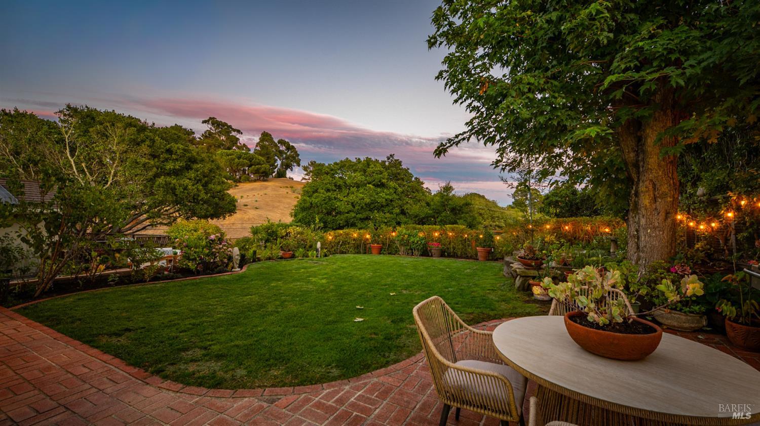 Landscaped backyard with a broad lawn, brick patio, outdoor dining table, and hillside view at dusk.