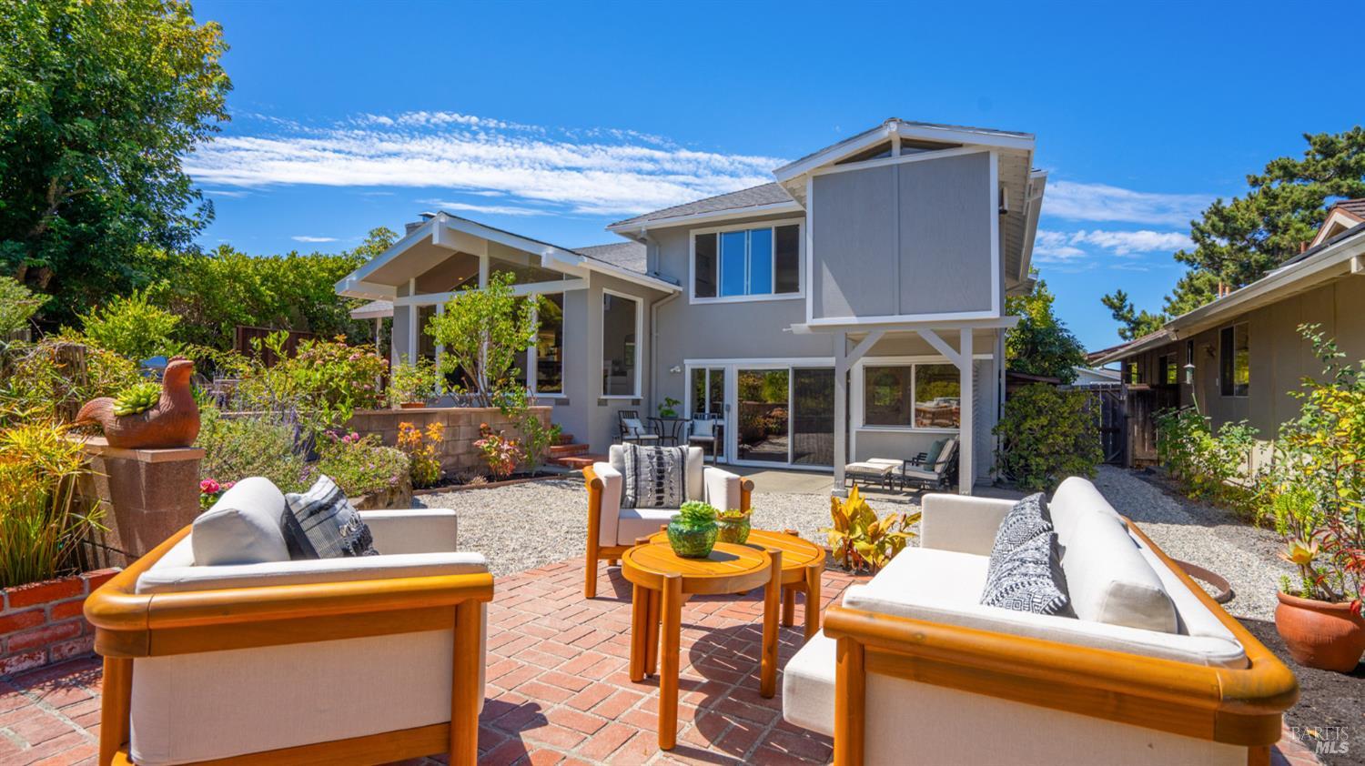Modern gray home behind a brick patio with white-cushioned seating and colorful garden beds.