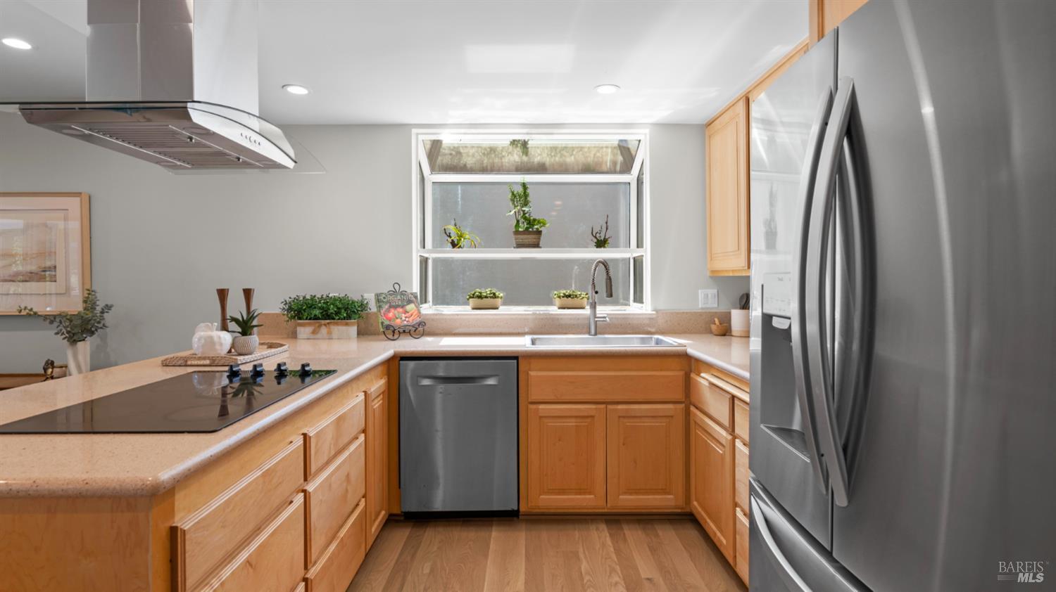 Kitchen with honey-colored wood cabinets, pale countertops, stainless steel appliances, and a wide window above the sink.