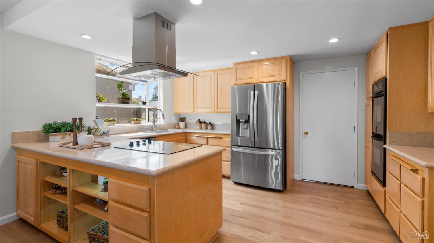 Kitchen with a large cooktop island, light wood cabinets, and stainless steel appliances.