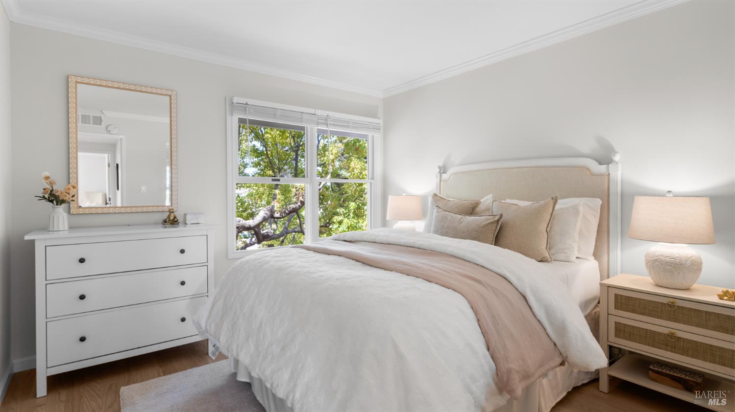 Furnished bedroom with a white bed and leafy trees visible through the window.