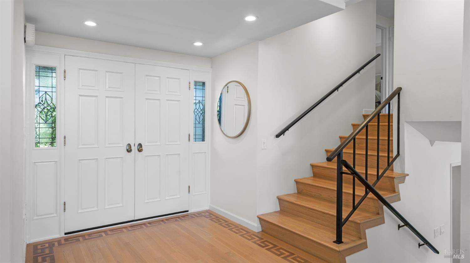 Entry foyer with stained-glass sidelights, patterned hardwood flooring, and a wood staircase with black railings.