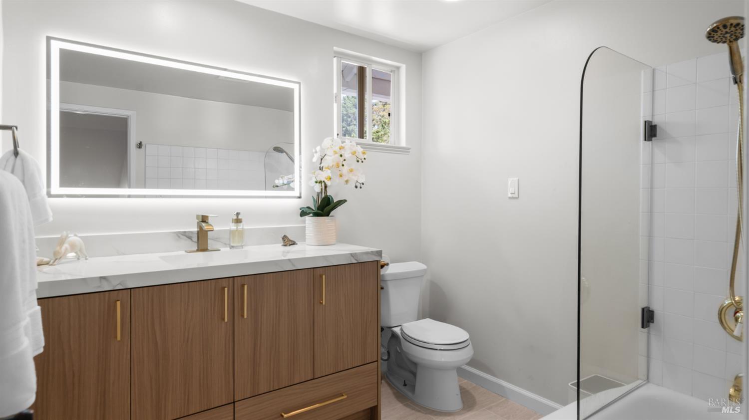 Bright bathroom with a wood vanity, illuminated mirror, and glass-enclosed shower.