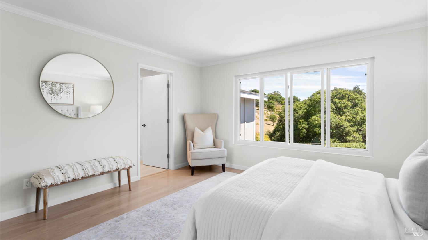 Bright bedroom with a white bed, round mirror, and wide windows overlooking trees.