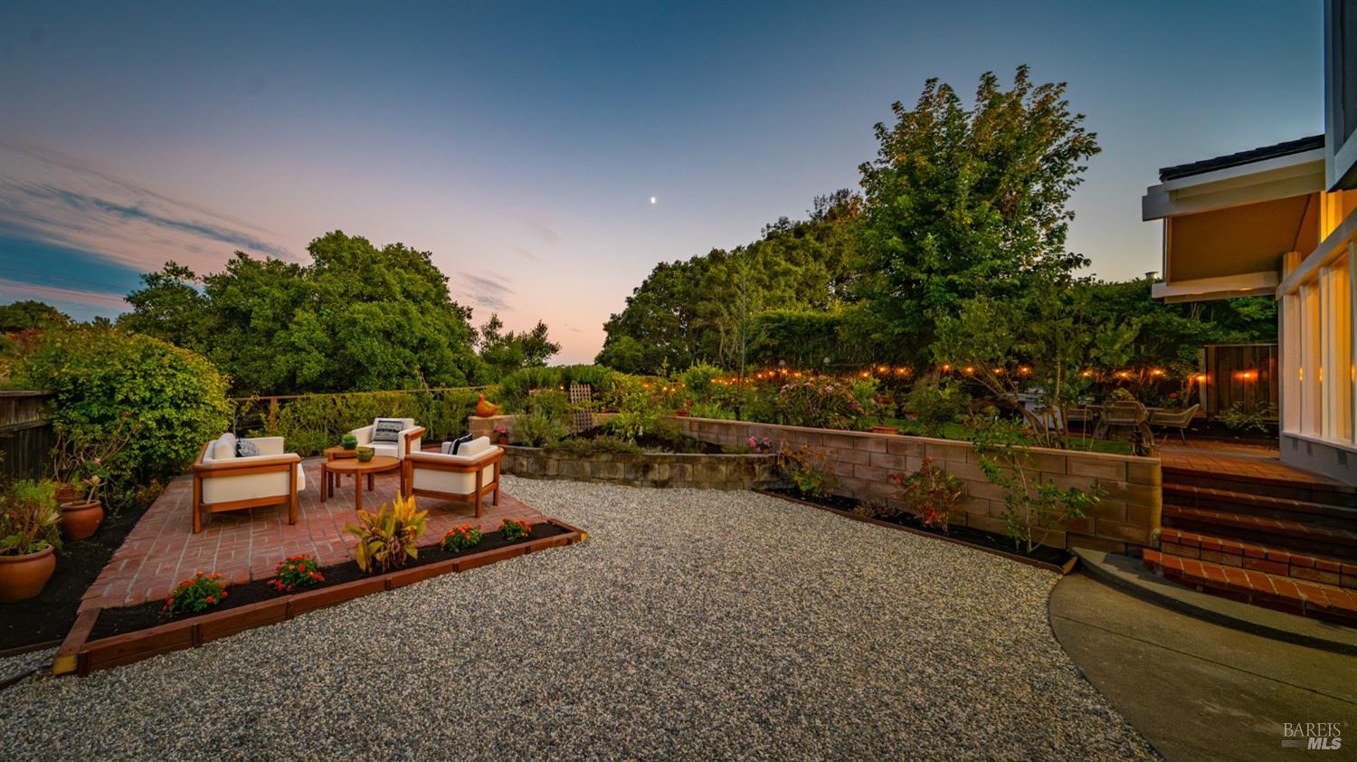 Landscaped backyard at dusk with a gravel courtyard, brick patio seating, raised beds, and glowing string lights.