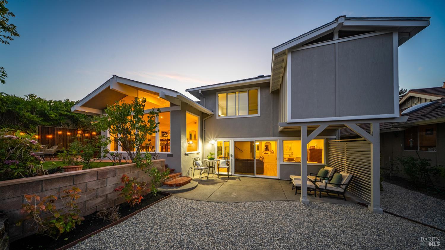 Modern two-story home at dusk, with a warmly lit covered patio and outdoor seating.
