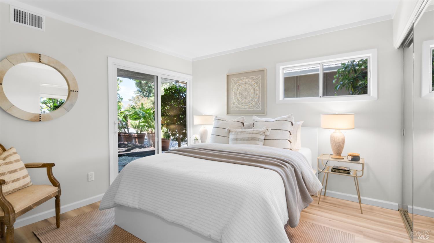 Staged bedroom with a white bed, round mirror, vintage chair, and sliding doors to a leafy patio.