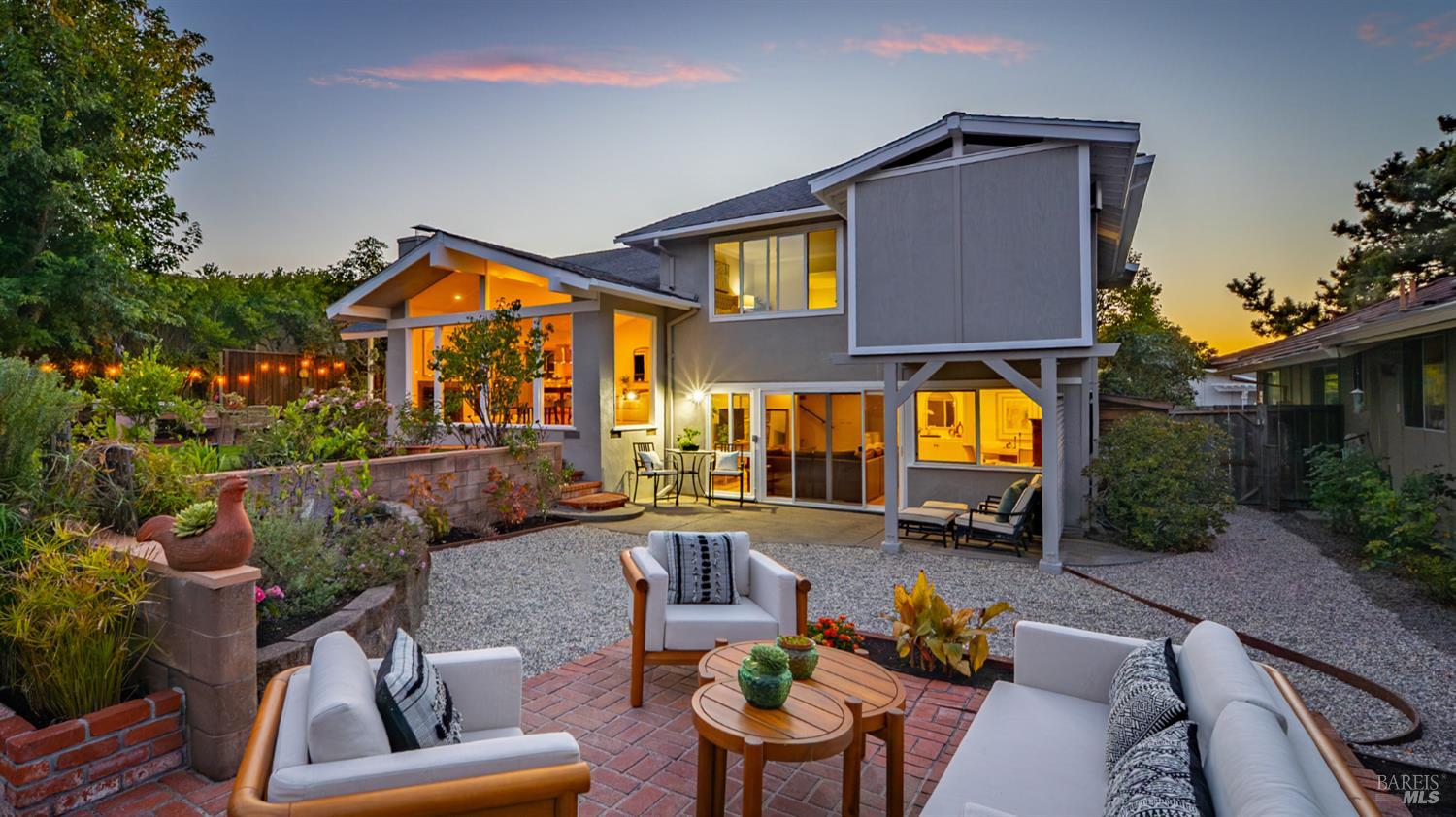 Modern gray home with glowing windows behind a furnished brick patio and landscaped gravel yard at dusk.
