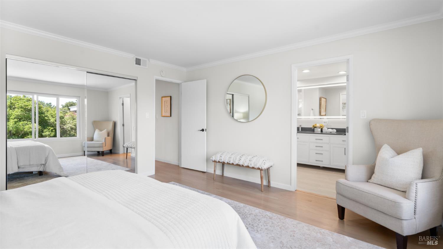 Bright bedroom with mirrored closet doors, a round wall mirror, and an open doorway to the bathroom.