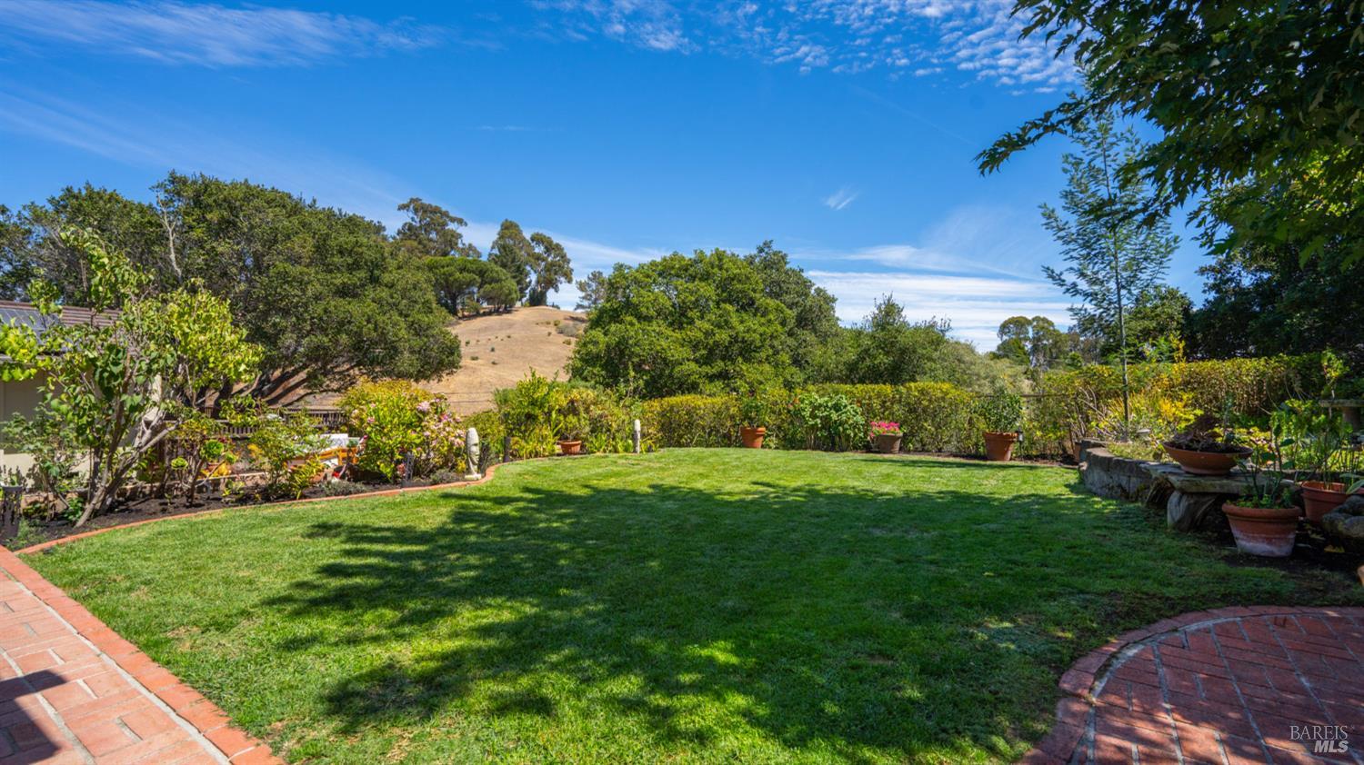 Landscaped backyard with a broad lawn, brick patio edging, and a tree-dotted hillside beyond.