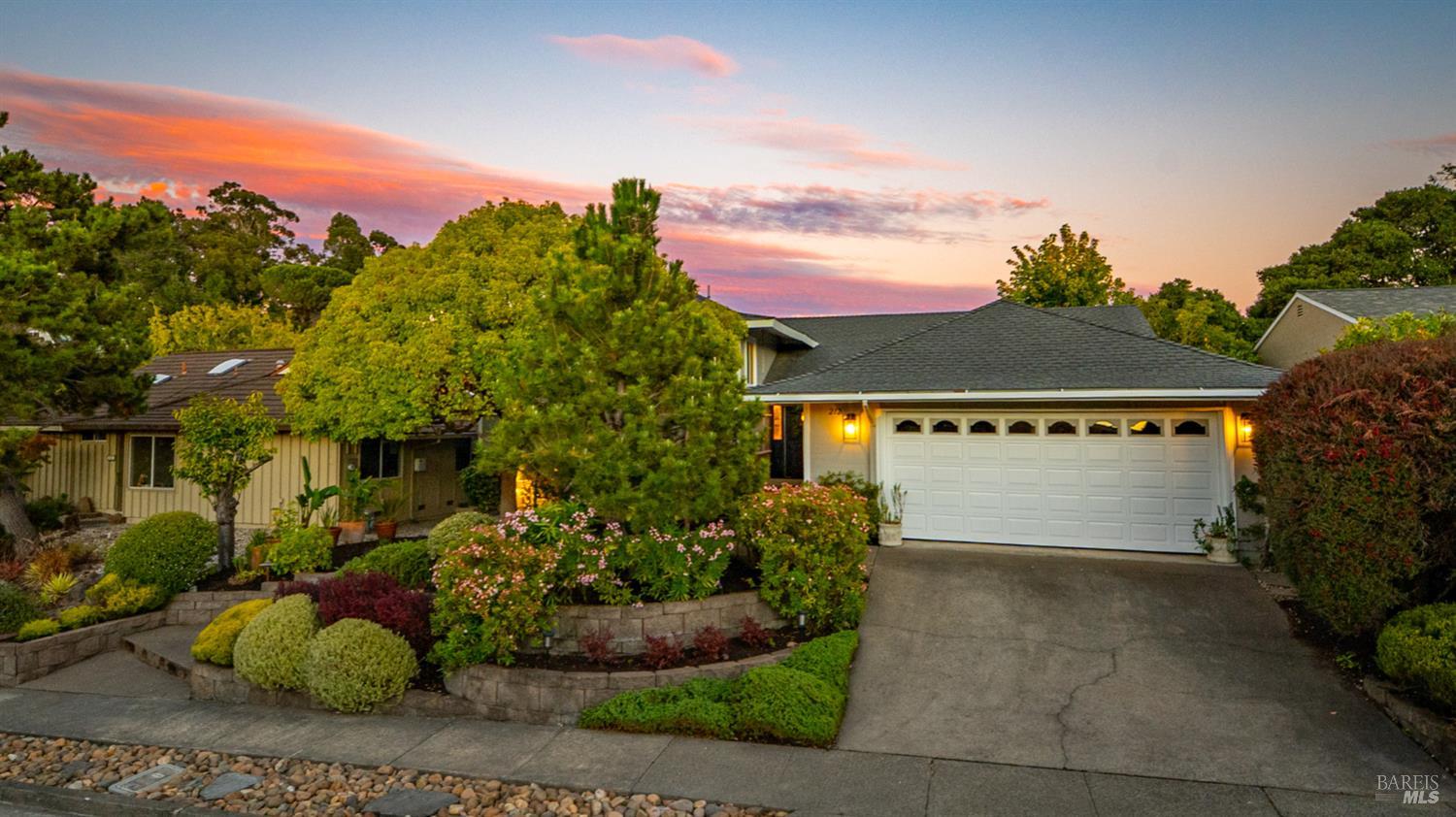 Single-story home with a white double garage, wide driveway, and landscaped flowering garden at dusk.