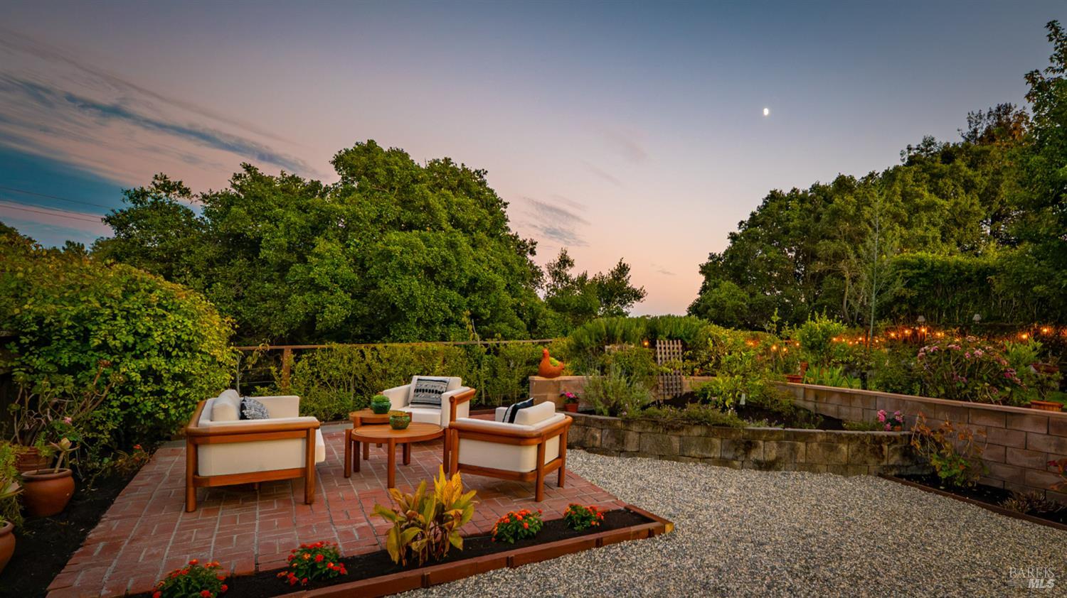 Backyard patio with cushioned lounge chairs, raised garden beds, and string lights at dusk.