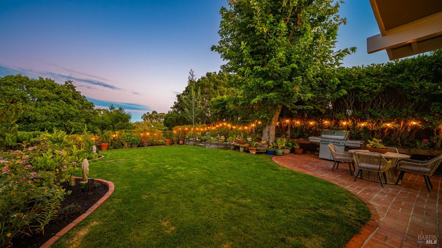 Landscaped backyard with a broad lawn, brick patio, outdoor dining set, and built-in grill at dusk.