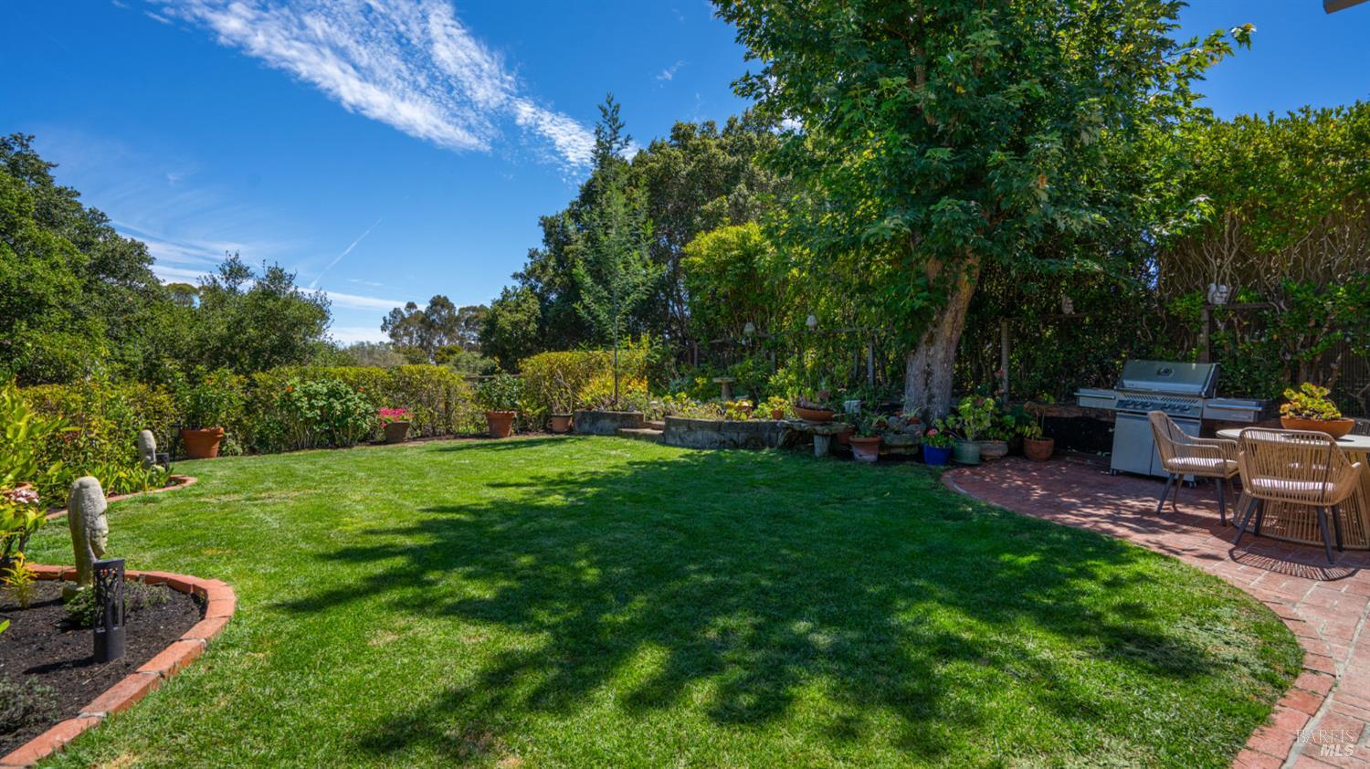 Landscaped backyard with a broad lawn, mature shade trees, and a brick patio with dining furniture and grill.