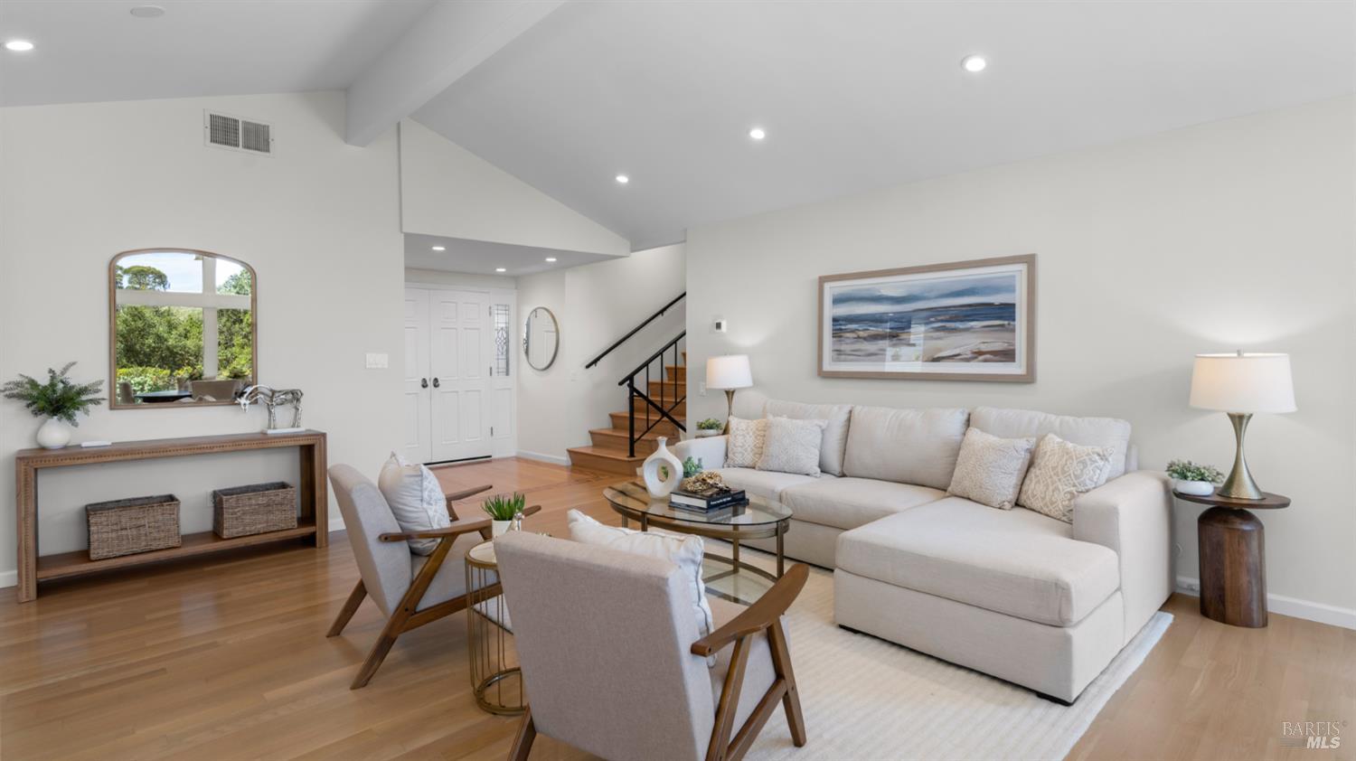 Staged living room with a white sectional, wood-framed chairs, and a staircase.