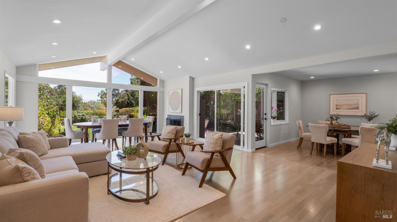 Open-plan living and dining room with beige seating, light wood floors, and large garden-facing windows.