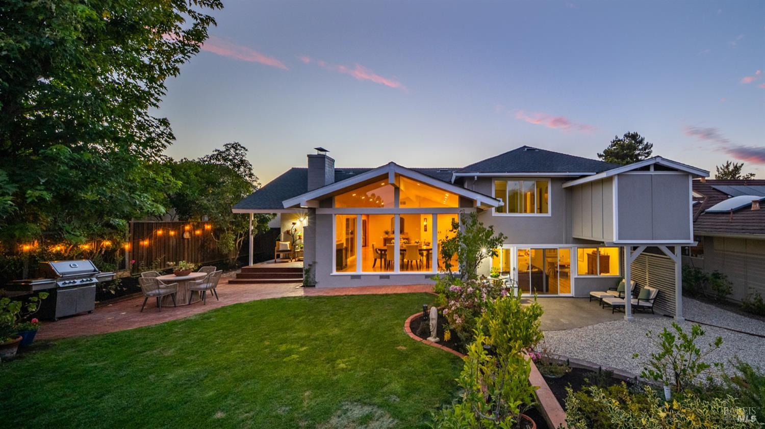 Modern two-story home with a lit glass facade, landscaped lawn, and brick patio at dusk.