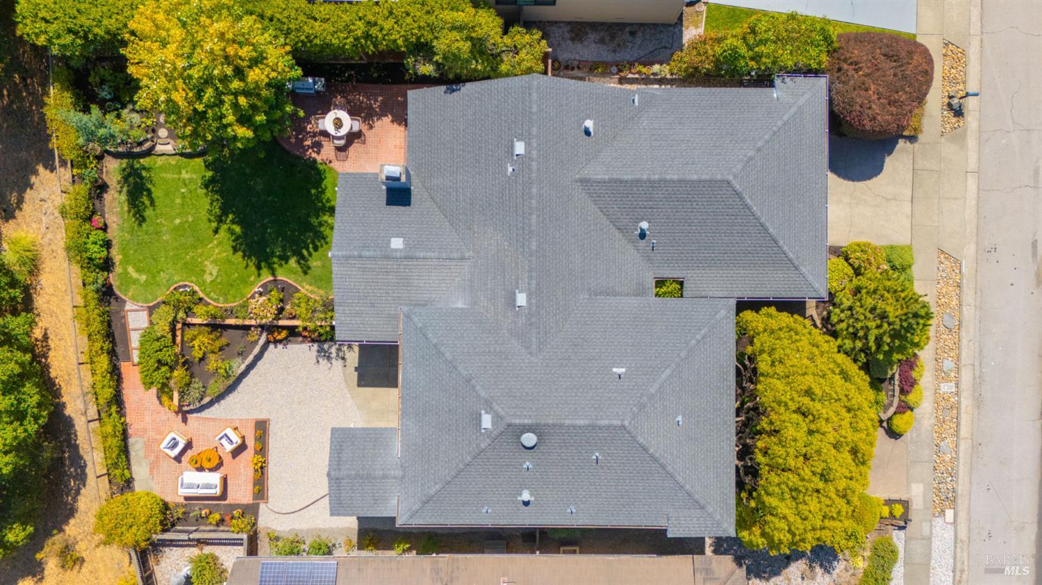 Aerial view of a gray-roofed home with landscaped gardens, a lawn, and brick patios.