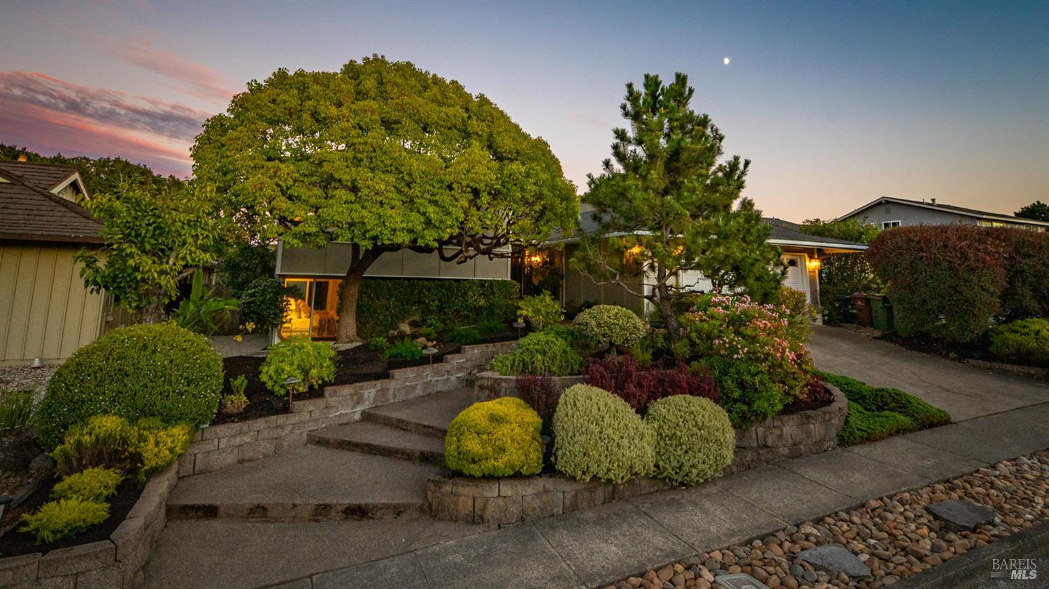 Single-story home framed by mature trees above a landscaped front garden and stone retaining walls at dusk.