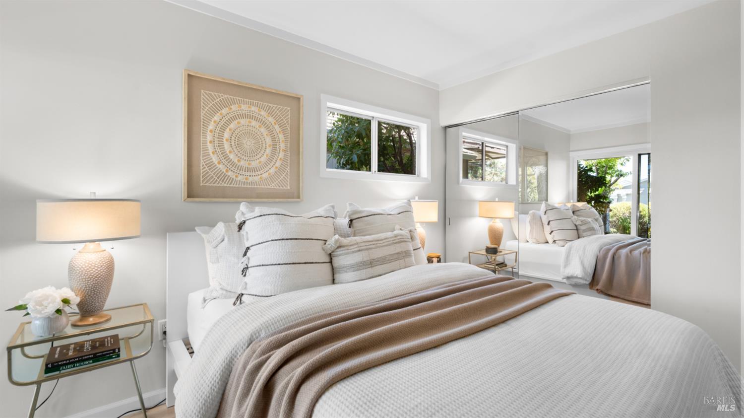 Staged bedroom with a white bed, taupe throw, framed circular artwork, and mirrored closet doors.
