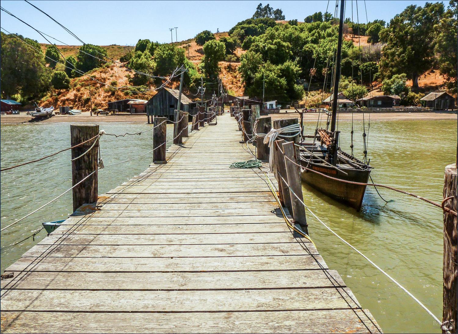 Wooden dock extends over water toward shoreline buildings and dry, tree-covered hills.