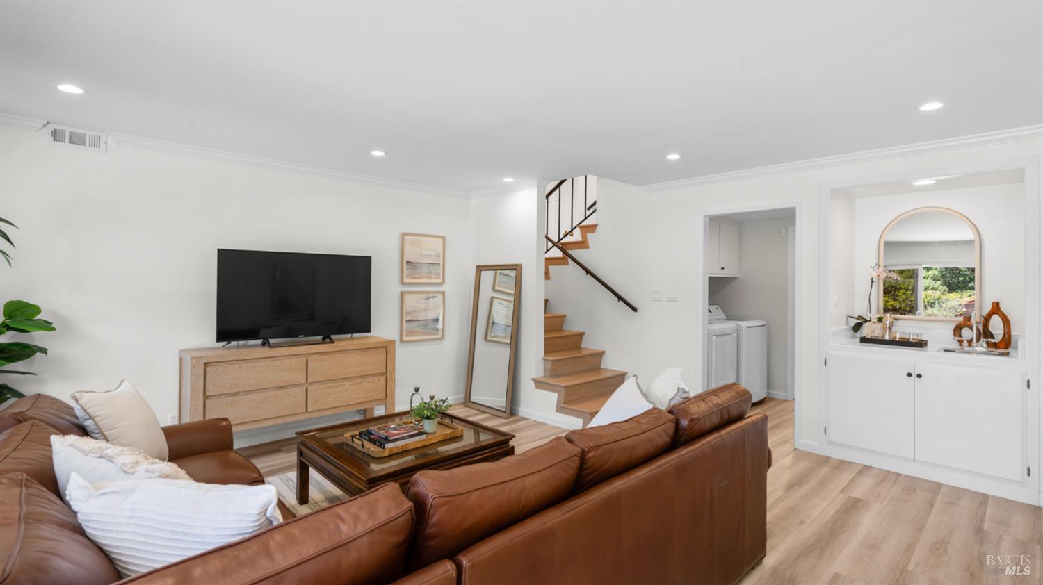Living room with a brown leather sectional, light wood floors, and an open staircase.