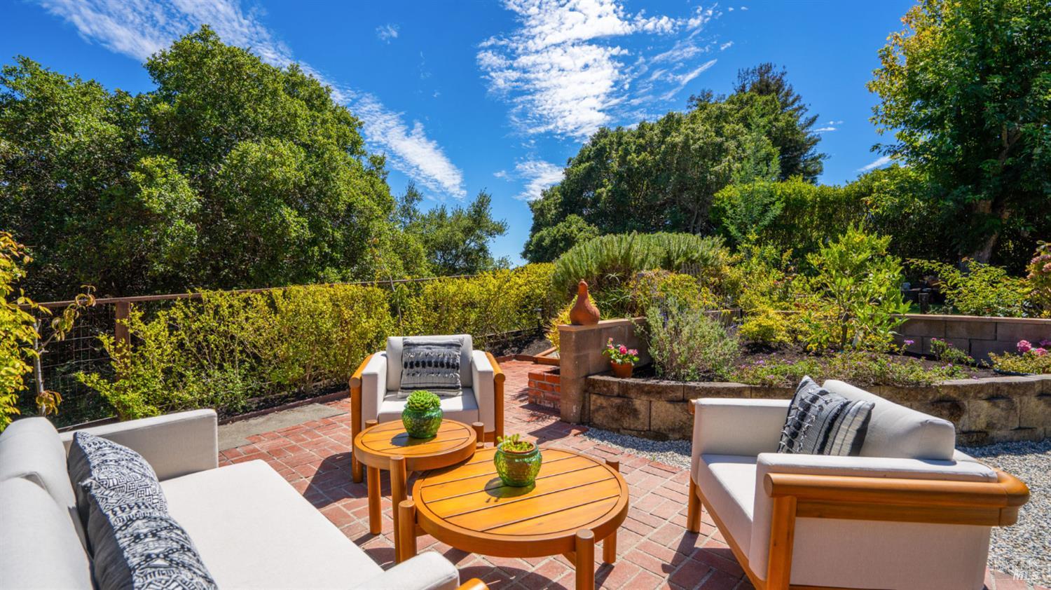 Brick patio with cushioned outdoor seating and round wooden tables, surrounded by lush garden beds.