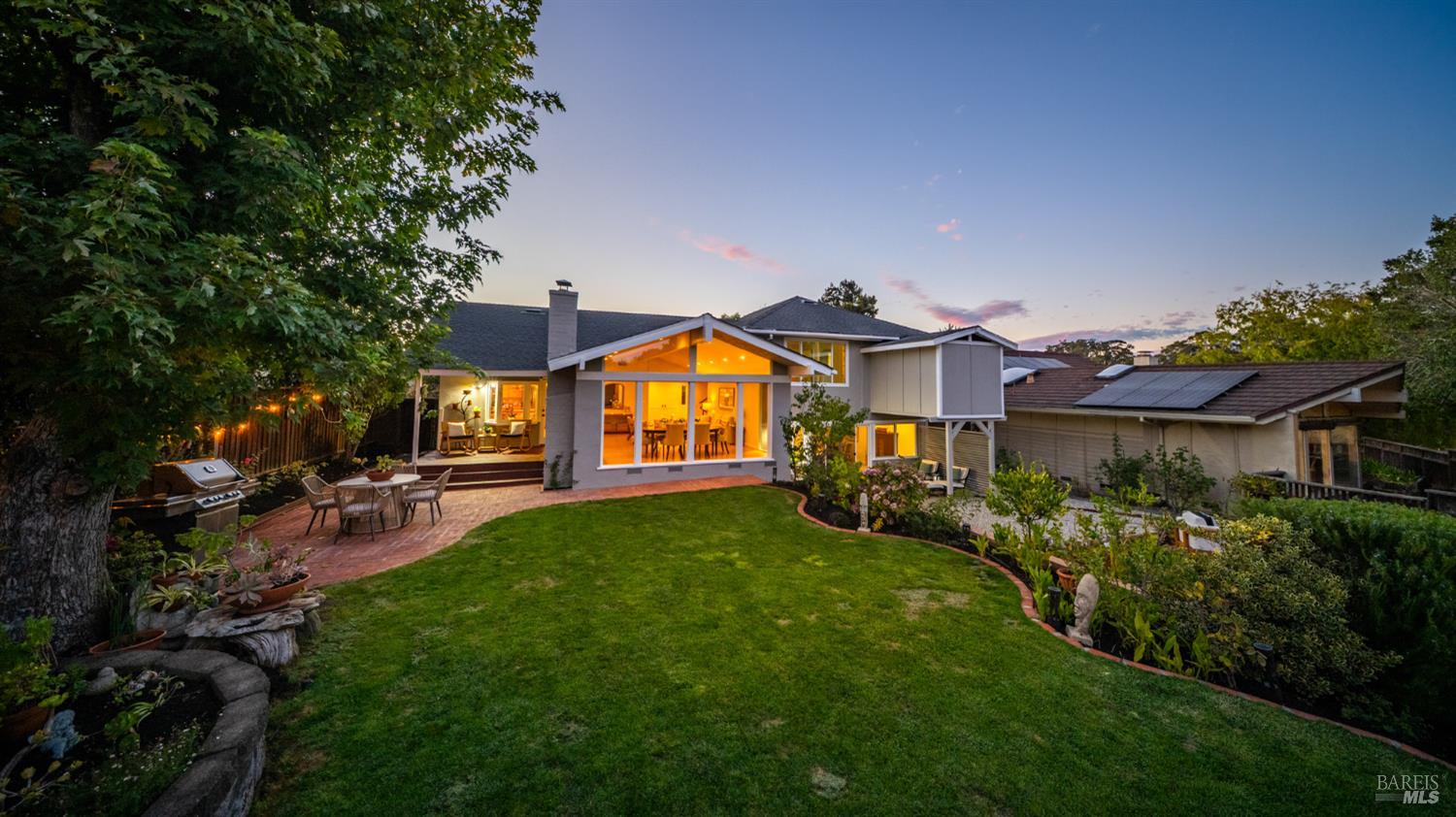 Home’s rear exterior at dusk, with a broad lawn, brick patio, glowing windows, and rooftop solar panels.