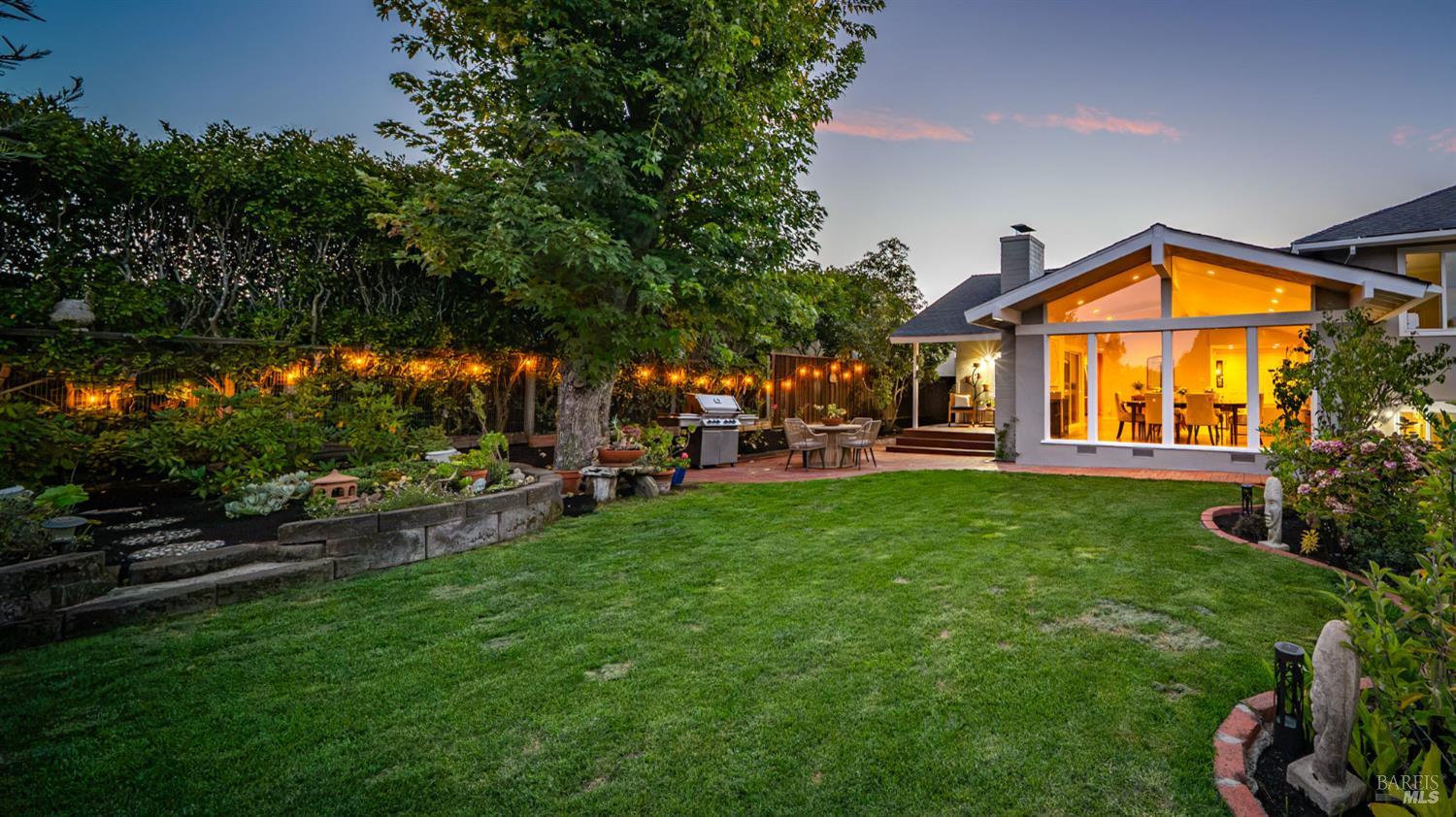 Home's backyard at dusk, with a broad lawn, raised garden beds, and a glowing dining room.