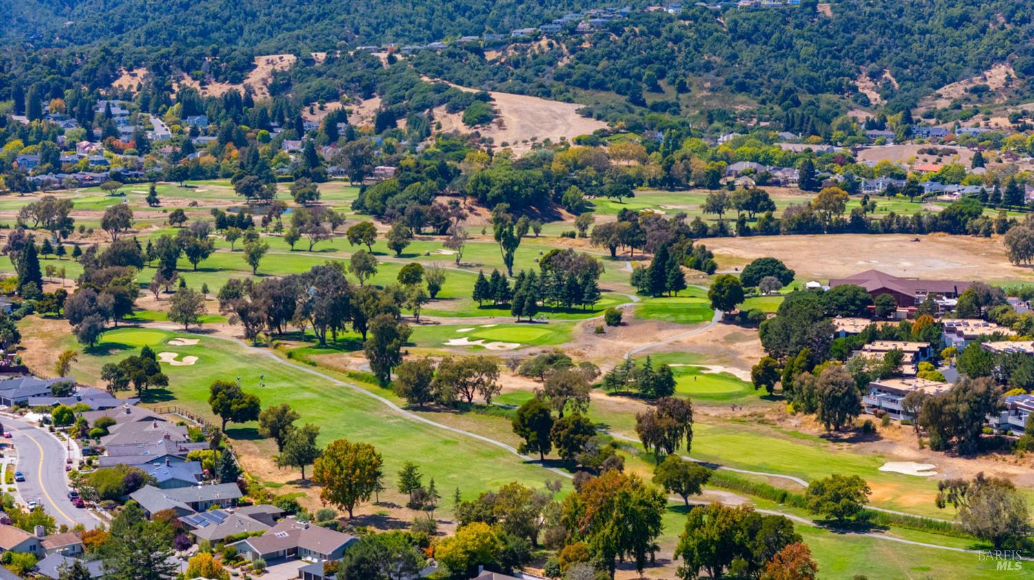 Aerial view of a golf course with sand bunkers, homes, and wooded hills beyond.
