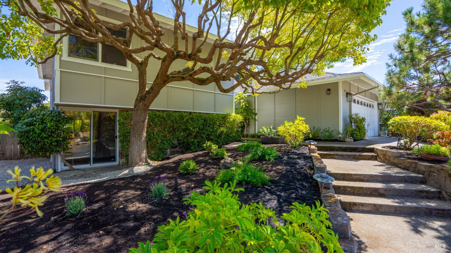 Two-story home with pale siding, landscaped garden, garage, and steps beneath a mature shade tree.