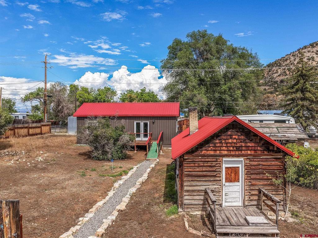 Rustic log cabin with a red roof beside a gravel path, with a second red-roofed building behind it.