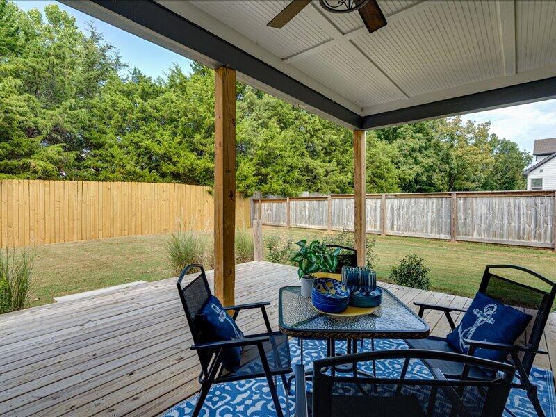 Covered wooden deck with a patio dining set and blue rug overlooking a fenced, tree-lined yard.