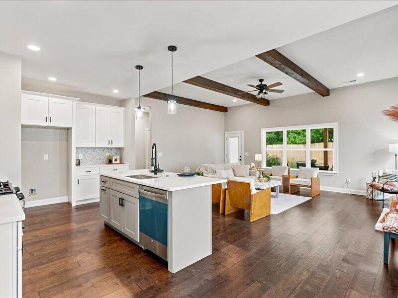 Open-plan kitchen and living area with a white island, exposed wood beams, and seating beside a large window.