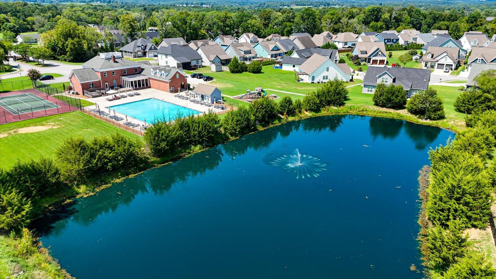 Aerial view of a neighborhood pond with a fountain, community pool, and tennis courts.