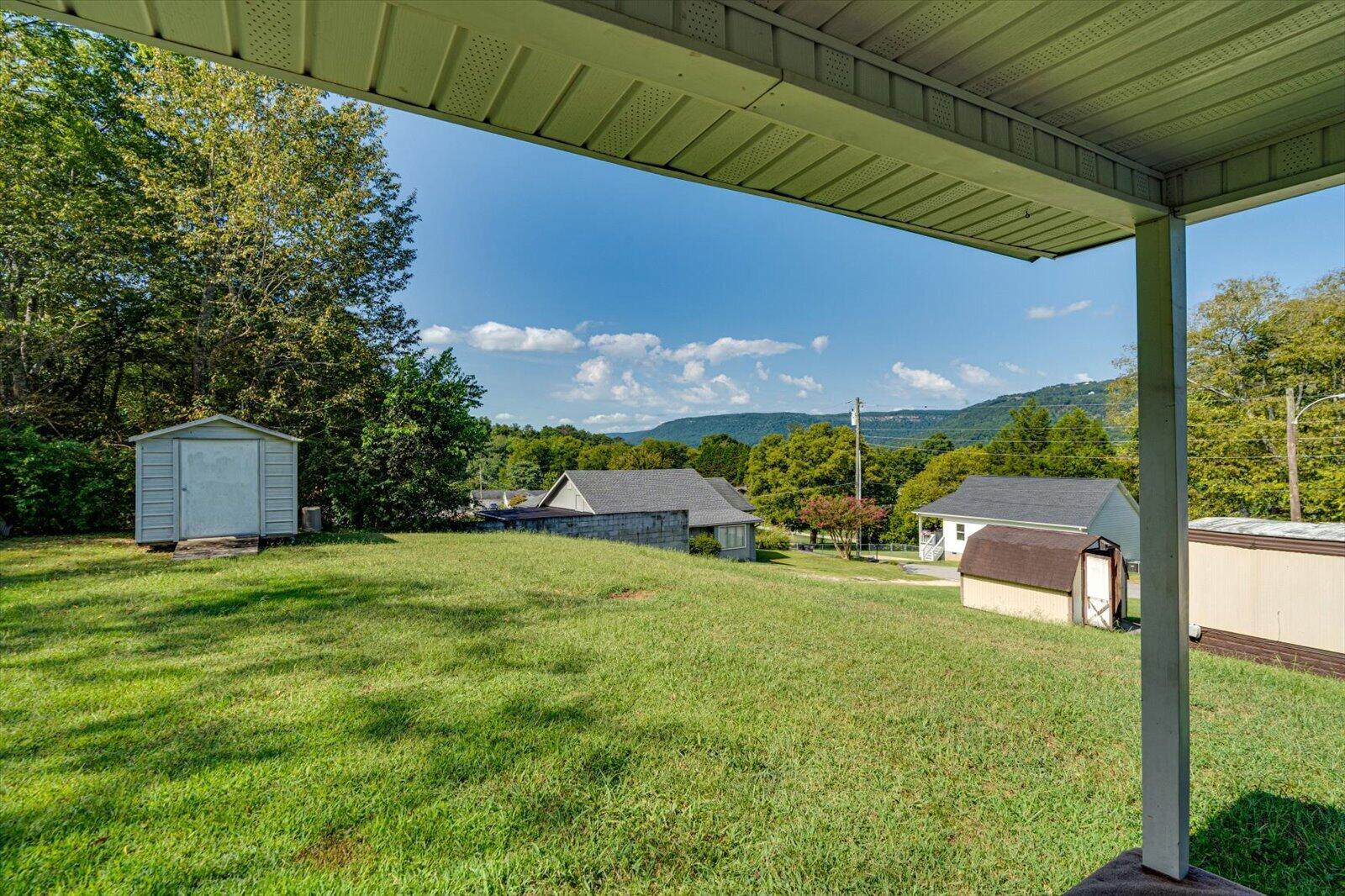Grassy backyard with sheds and distant wooded hills, viewed from beneath a covered porch.